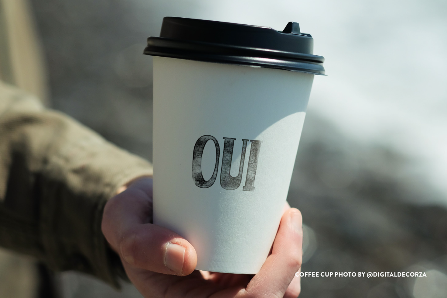 Hand holding a paper coffee cup with "OUI" text for mockup, clear typeface, ideal for designers to showcase branding designs.